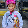 bokeh, car, cartoon_dog, child, clothing, crown, handmade_crown, happy, hearts, outdoor, parking_lot, pavement, pink, portrait, sequins, smile, sweatshirt, toddler, wheel, young_child