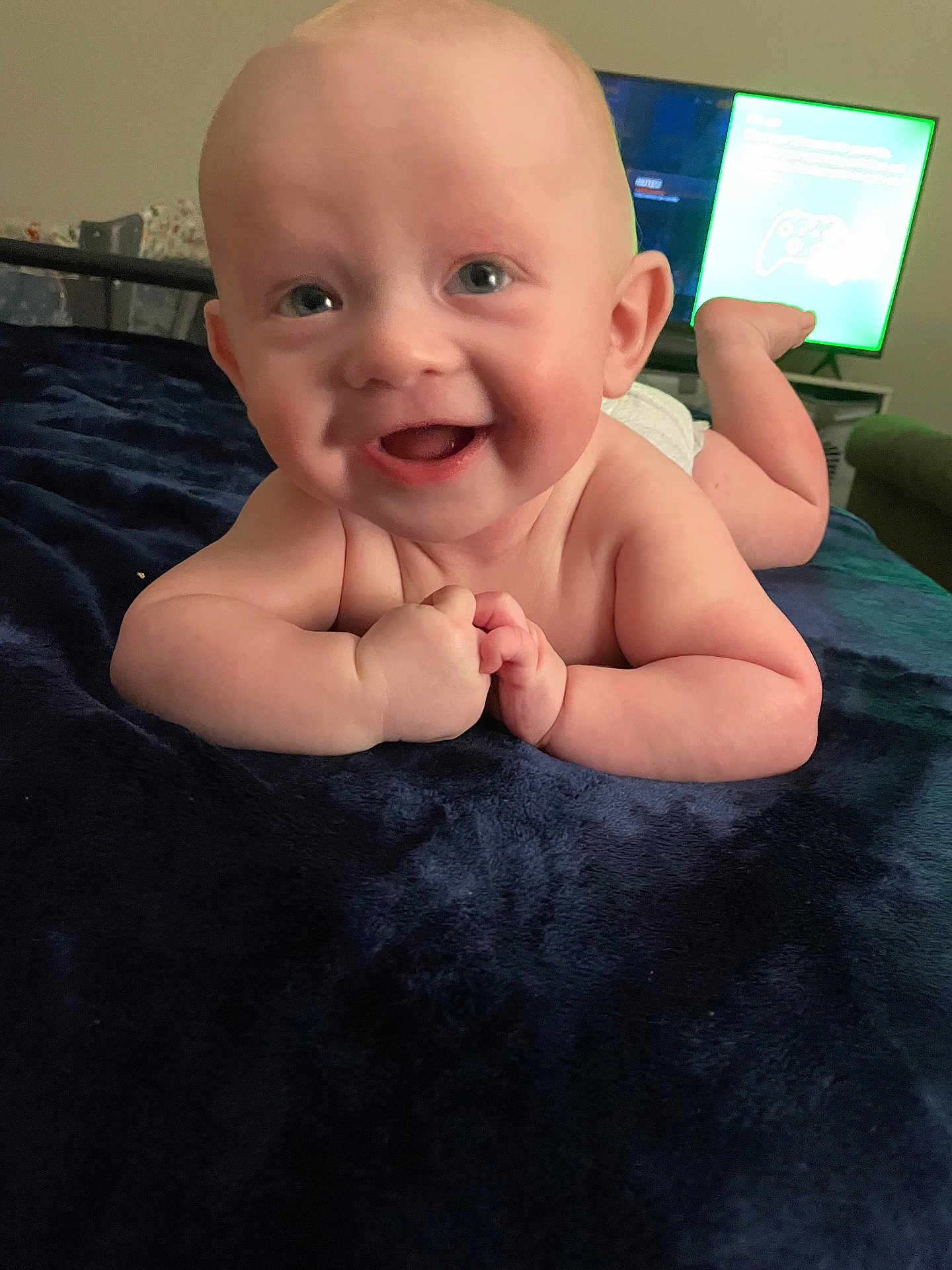 Bryson is registered to the contest to win money with this photo: baby, smiling, infant, blanket, blue_blanket, face, hands, happy, lying_down, indoor, television, living_room, skin, cute, child, young, person, expression, home, furniture