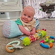 Connor is registered to the contest to win money with this photo: baby, infant, toy, carpet, pillow, smile, face, indoor, child, playtime, colorful, cute, person, floor, baby_toy, nursery, soft_lighting, happy, bald, clothing