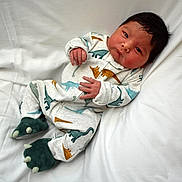 Esteban is registered to the contest to win money with this photo: baby, infant, dinosaur_onesie, clothing, footwear, slippers, soft_surface, white_background, lying_down, cute, child, newborn, indoors, portrait, face, hands, expression, cozy, sleepwear, resting