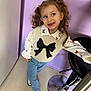 child, girl, smiling, curly_hair, pigtails, bow, sweater_vest, butterfly_pattern, shirt, jeans, silver_shoes, barstool, purple_wall, indoor, floor, playful, happy, standing, looking_away, person