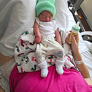 Archie is registered to the contest to win money with this photo: newborn, baby, infant, hat, knit_hat, bottle, blanket, footprint_pattern, hospital, person, hand, clothing, socks, bed, resting, indoor, pink_shirt, white_shirt, baby_clothes, closeup