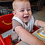 Henry is registered to the contest to win money with this photo: baby, child, high_chair, smiling, happy, indoor, table, red_chair, clothing, bib, face, person, wooden_chair, floor, cafe, furniture, arms, skin, expression, tabletop