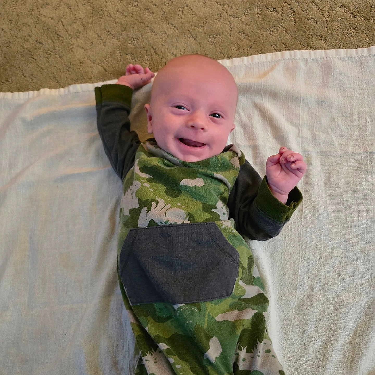 Travis is registered to the contest to win money with this photo: baby, infant, child, smiling, lying_down, portrait, face, camouflage, pajamas, blanket, bed, indoor, happy, arms_raised, fist, bald, looking_at_camera, close_up, cute, photography