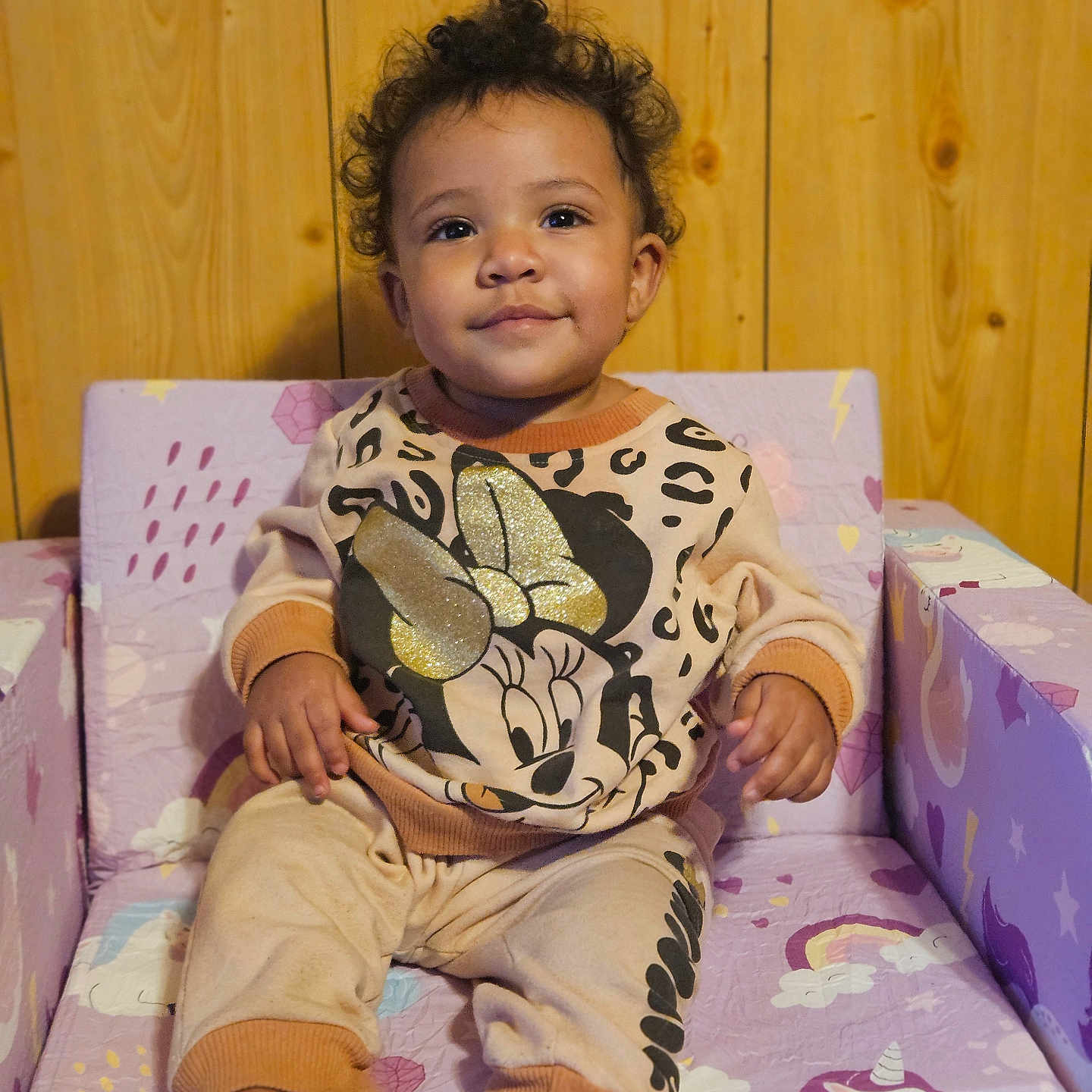 Xana is registered to the contest to win money with this photo: barefoot, chair, child, comfortable, cozy, curly_hair, cute, glitter, indoors, minnie_mouse, pants, pink, portrait, purple, sitting, smiling, sweatshirt, toddler, unicorn, wooden_wall