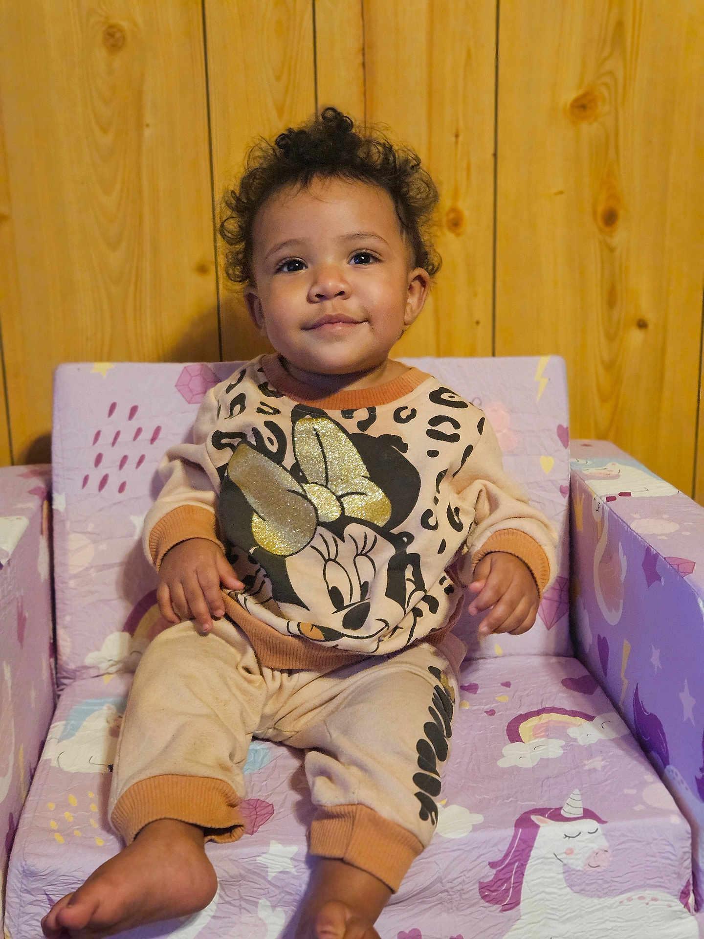 Xana is registered to the contest to win money with this photo: toddler, child, smiling, curly_hair, sitting, minnie_mouse, sweatshirt, pants, barefoot, unicorn, chair, purple, pink, wooden_wall, indoors, cozy, cute, comfortable, glitter, portrait