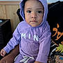 Xana is registered to the contest to win money with this photo: child, toddler, hoodie, purple_clothing, indoor, person, adult_hand, face, portrait, seated, curious_eyes, casual_wear, wood_paneling, wall, graphic_tshirt, animation_print, home, expression, baby, closeup