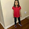 child, girl, standing, portrait, indoors, hallway, red_shirt, black_pants, sneakers, long_hair, smiling, face, wood_floor, wall, outlet, painting, home, casual_clothing, full_body, photograph