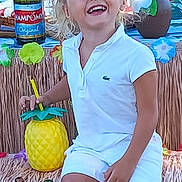 Lana participe au concours pour gagner de l'argent avec cette photo : child, smiling, curly_hair, white_clothing, pineapple_cup, tiki_theme, party, decorations, colorful, happy, sitting, drink, table, bottle, people, festival, tropical, celebration, fun, outdoor