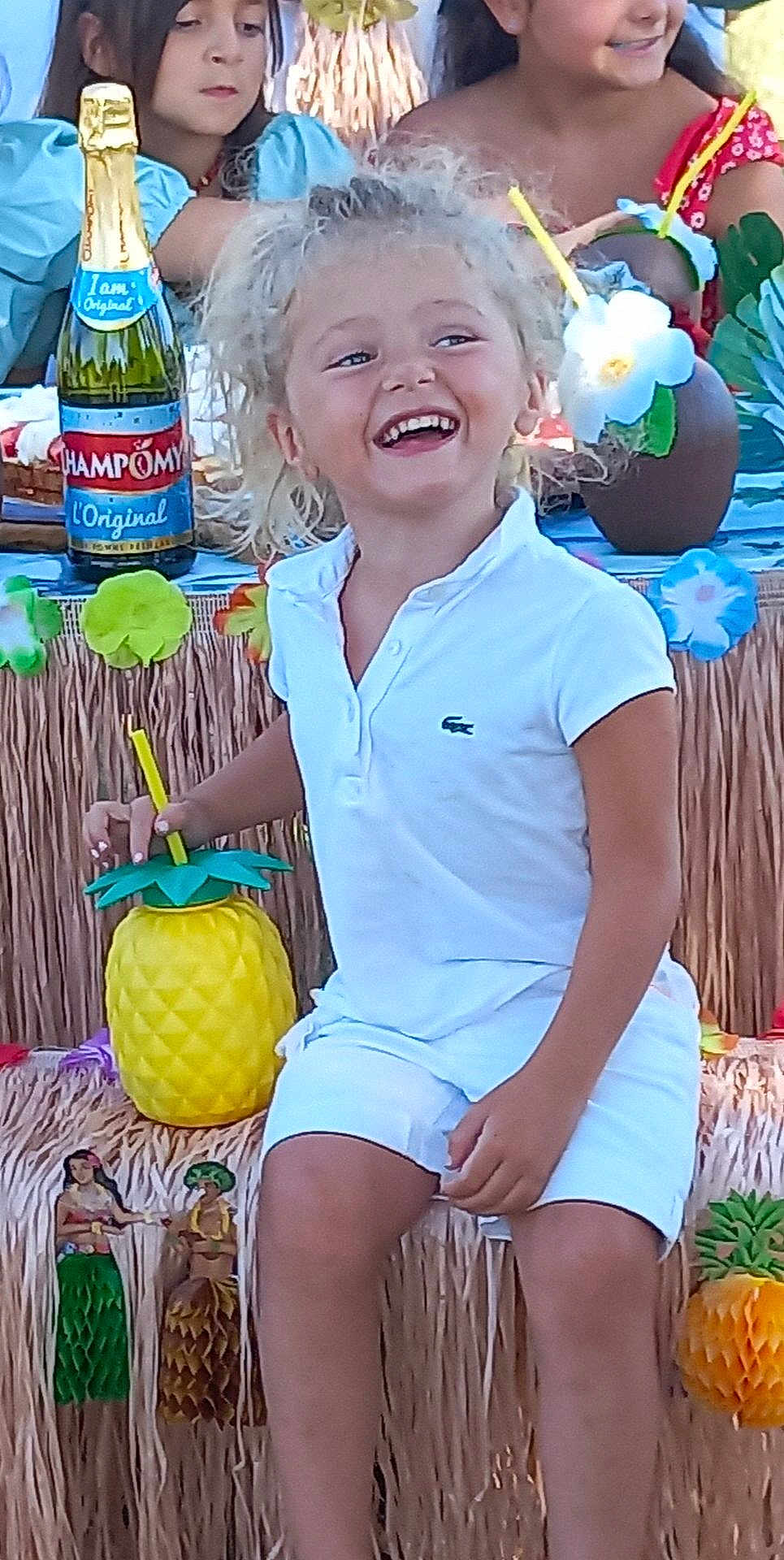 Lana participe au concours pour gagner de l'argent avec cette photo : child, smiling, curly_hair, white_clothing, pineapple_cup, tiki_theme, party, decorations, colorful, happy, sitting, drink, table, bottle, people, festival, tropical, celebration, fun, outdoor