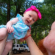 Ensley is registered to the contest to win money with this photo: arm, baby, blue_dress, child, daylight, flamingo_pattern, forest, happy, holding, nature, outdoor, parked_cars, person, pink_headband, playful, portrait, red_beard, smiling, tattoo, trees