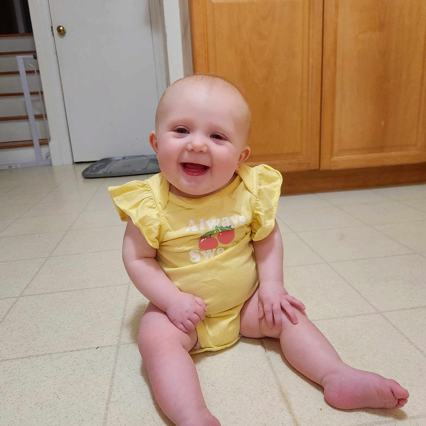 Faye is registered to the contest to win money with this photo: baby, bright, casual, child, chubby_cheeks, cute, door, floor, happy, home, indoors, infant, kitchen, person, short_hair, sitting, smiling, tile_floor, wooden_cabinets, yellow_clothing