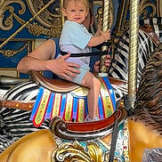 Everleigh joined the competition — help win amazing prizes! child, toddler, carousel, horse, adult, hands, ride, amusement_park, decorative, bright_colors, blue, gold, pattern, smile, shorts, shirt, footwear, fun, entertainment, family