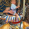 child, toddler, carousel, horse, adult, hands, ride, amusement_park, decorative, bright_colors, blue, gold, pattern, smile, shorts, shirt, footwear, fun, entertainment, family