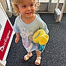 child, toddler, blonde_hair, curly_hair, blue_eyes, smiling, plush_toy, yellow_toy, elsa_shirt, sandals, indoor, carpet, store_entrance, door, sign, burlington, person, clothing, happy, cute