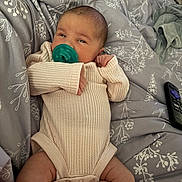 Kennedy is registered to the contest to win money with this photo: baby, pacifier, onesie, blanket, infant, lying_down, cute, face, hands, legs, pattern, fabric, bed, remote_control, soft, cozy, expression, skin, person, resting