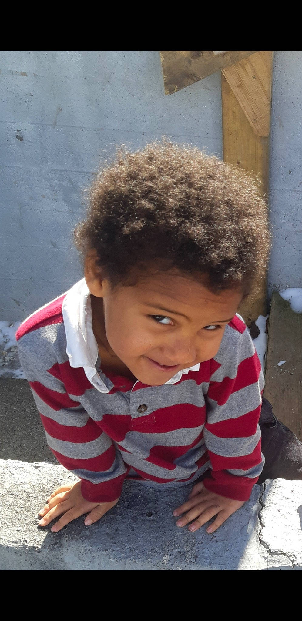 Lennon is registered to the contest to win money with this photo: afro, asphalt, chin, cool, eye, face, flooring, grass, happy, head, leisure, outerwear, person, plant, road_surface, sidewalk, sitting, skin, smile, toddler