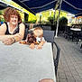 woman, child, toddler, stuffed_animal, plush_toy, table, outdoor, patio, awning, umbrella, chair, wicker_chair, hand, curly_hair, glasses, smile, person, restaurant, paving, garden