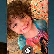 Harper is registered to the contest to win money with this photo: child, smiling, curly_hair, couch, blanket, colorful_clothing, portrait, indoor, phone_screen, face, happy, person, home, cozy, sitting, cute, young_child, digital_screen, app_icon, casual