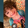 child, smiling, curly_hair, couch, blanket, colorful_clothing, portrait, indoor, phone_screen, face, happy, person, home, cozy, sitting, cute, young_child, digital_screen, app_icon, casual
