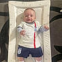 Ryder is registered to the contest to win money with this photo: baby, infant, smiling, england, soccer_outfit, changing_mat, star_pattern, white_clothing, blue_shorts, socks, nike, carpet, floor, indoor, child, cute, young, person, happy, portrait