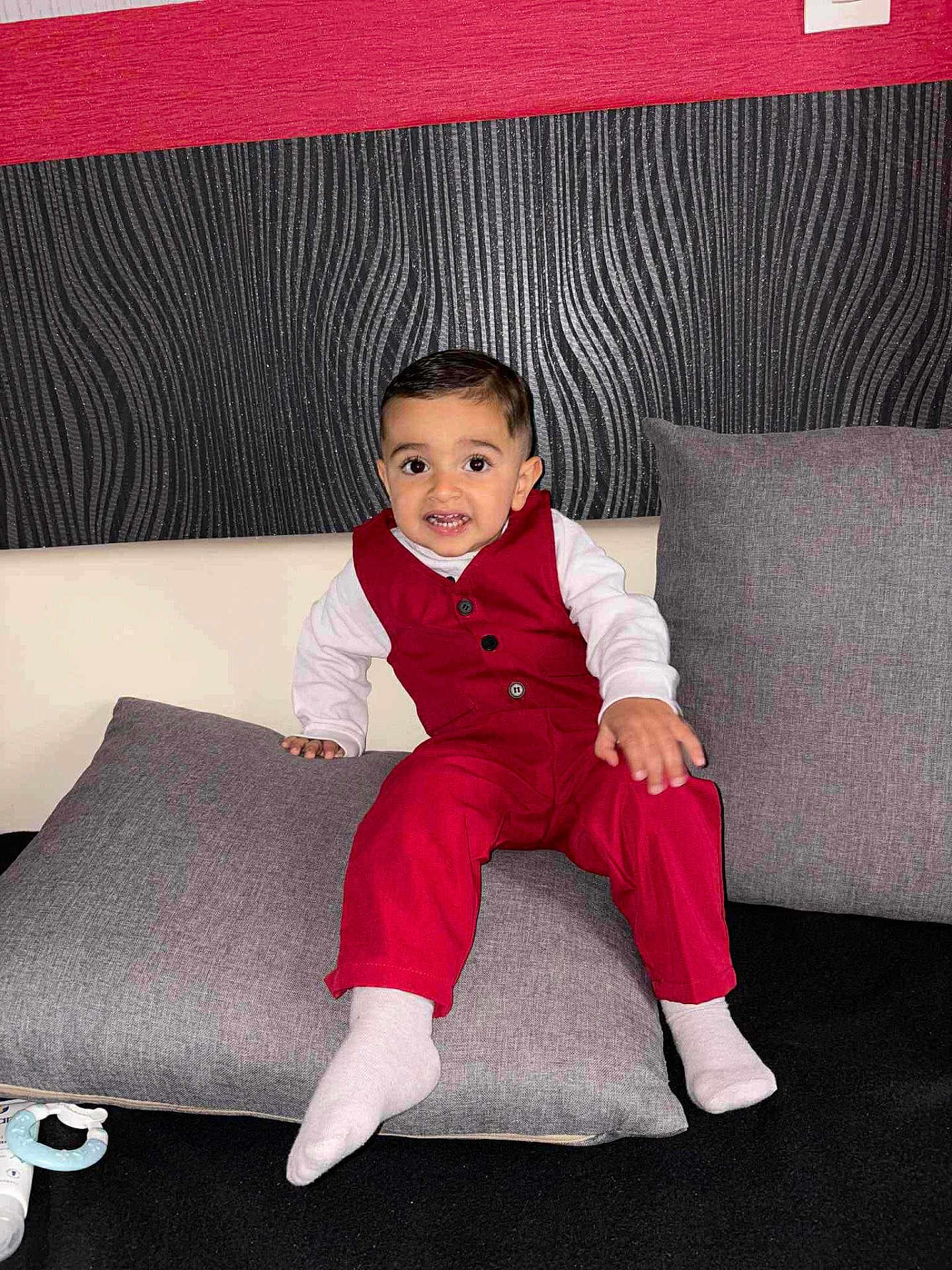 Alessyo a rejoint le concours — aidez-le/la à gagner de superbes lots ! toddler, child, red_outfit, cushion, gray_pillow, indoor, smiling, face, person, baby_toy, white_socks, seated, black_background, textured_wall, home, cute, fashion, portrait, young_child, furniture