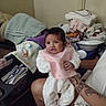 Donna is registered to the contest to win money with this photo: baby, child, infant, portrait, sitting, pink_bib, white_clothing, smiling, curious, face, indoors, laundry, folded_clothes, baby_gear, adult_arm, tattoo, home, chair, blanket, photography