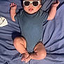 Malik is registered to the contest to win money with this photo: baby, infant, sunglasses, onesie, blue_bedspread, lying_down, tongue_out, cute, child, skin, face, legs, arms, hand, feet, relaxed, indoors, person, sleepwear, funny_expression