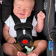 Malik is registered to the contest to win money with this photo: baby, car_seat, smile, infant, child, pacifier, seatbelt, clothing, white_shirt, brown_shorts, barefoot, cute, happy, safety, straps, indoor, person, young_child, seat, comfort