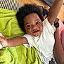 toddler, child, smiling, happy, arms_up, curly_hair, white_shirt, green_blanket, striped_couch, wooden_floor, indoor, person, baby, cute, playful, face, skin, expression, joy, home