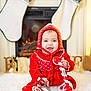 baby, child, red_clothing, knitwear, hat, smiling, telephone, toy, stockings, fireplace, holiday, christmas, decorations, believe_sign, white_rug, indoors, happy, festive, cute, portrait