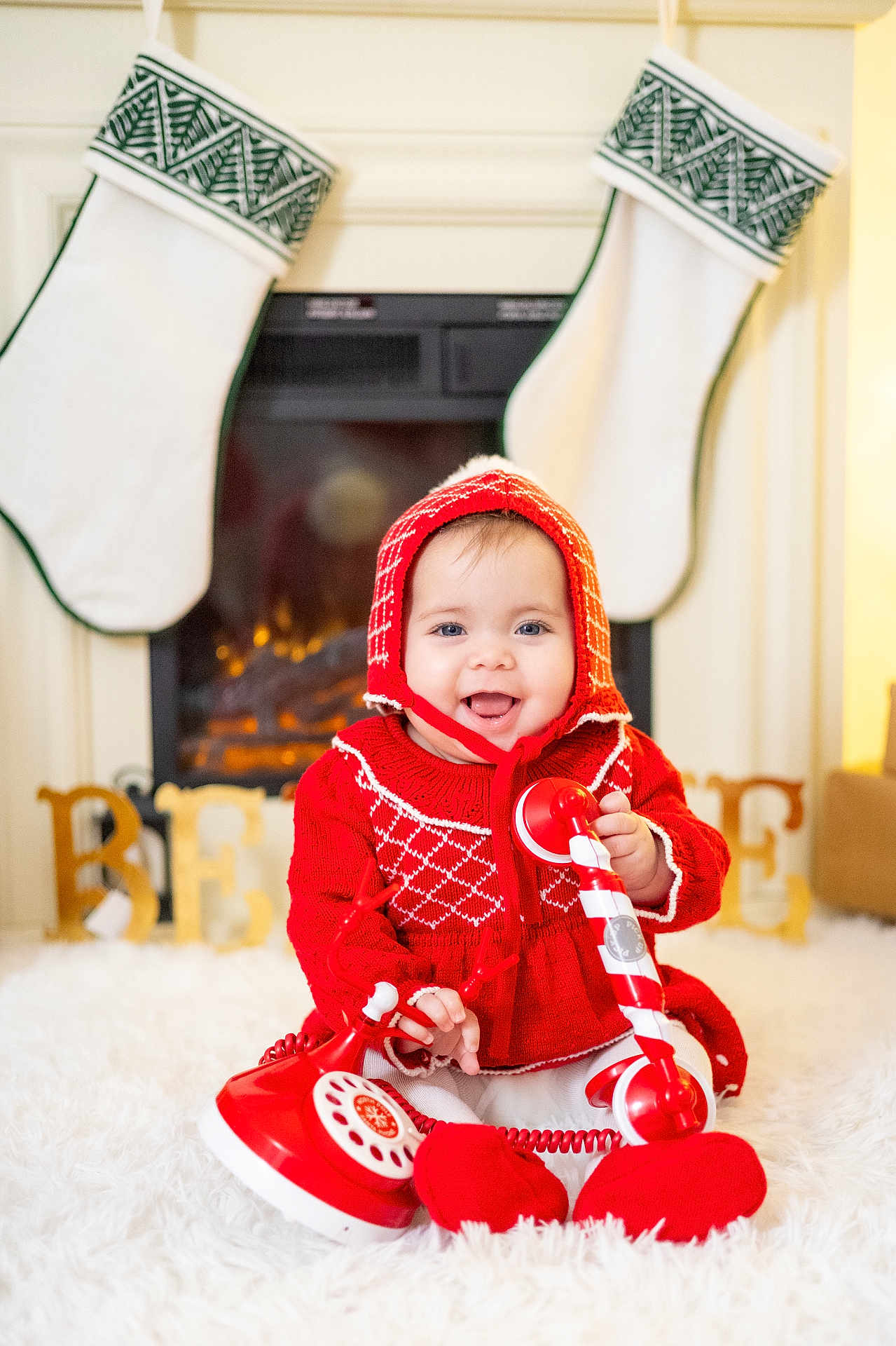 Eloa is registered to the contest to win money with this photo: baby, child, red_clothing, knitwear, hat, smiling, telephone, toy, stockings, fireplace, holiday, christmas, decorations, believe_sign, white_rug, indoors, happy, festive, cute, portrait