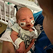 Remington is registered to the contest to win money with this photo: baby, infant, hospital, medical_tape, nasal_tube, crib, adult_hand, blue_clothing, white_shirt, holding, newborn, medical_bracelet, person, bedroom, healthcare, closeup, cute, care, tubes, patient