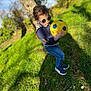 toddler, child, sunglasses, soccer_ball, grass, tree, sunlight, outdoor, jeans, sneakers, hoodie, nature, playing, daylight, casual_clothing, person, greenery, flower, park, cute