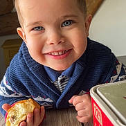 Keylan participe au concours pour gagner de l'argent avec cette photo : child, smiling, curly_hair, blue_eyes, sweater, indoor, wooden_table, pastry, food, hand, face, portrait, cozy, rustic, beam, cute, happy, person, closeup, breakfast