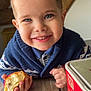 child, smiling, curly_hair, blue_eyes, sweater, indoor, wooden_table, pastry, food, hand, face, portrait, cozy, rustic, beam, cute, happy, person, closeup, breakfast
