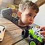 toddler, child, toy, monster_truck, table, wooden_table, striped_shirt, indoor, curious, hair, face, hand, room, wooden_beams, furniture, play, vehicle, focus, closeup, person