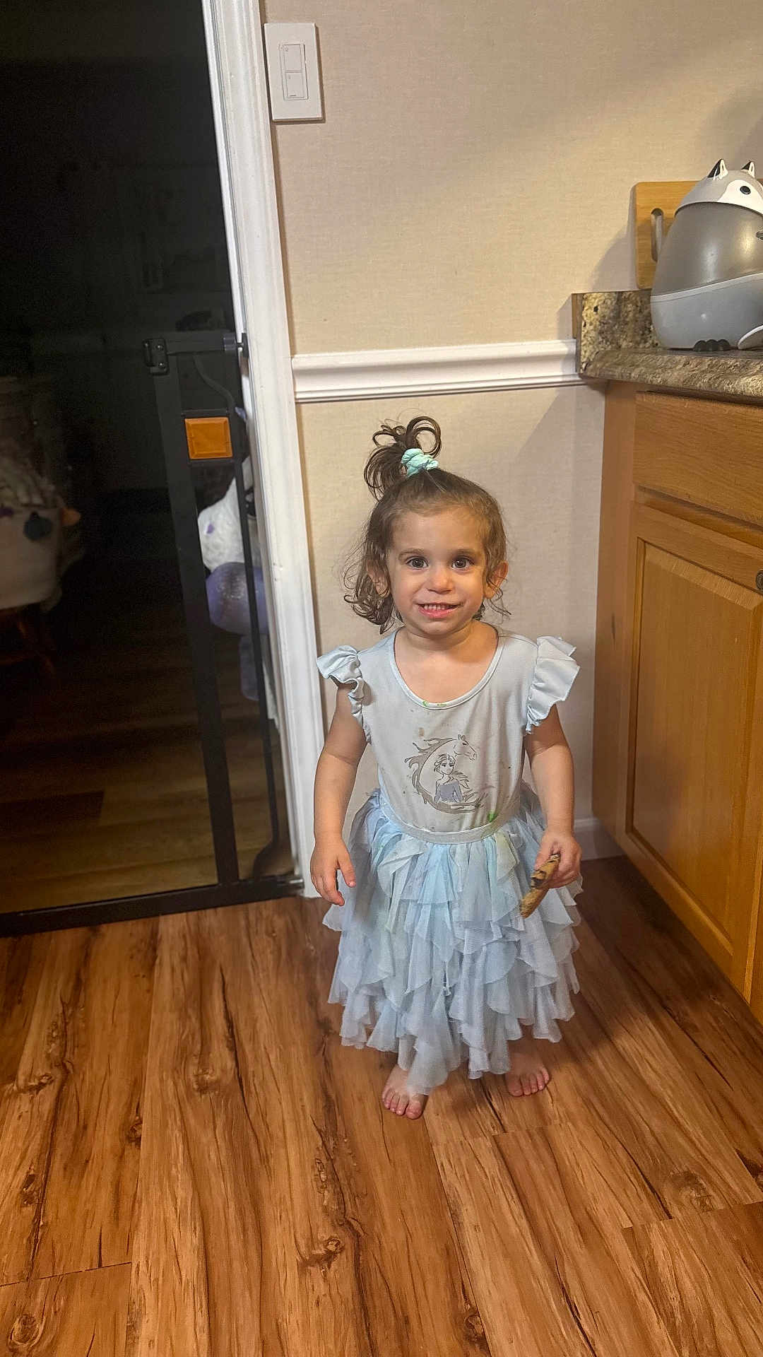 Colby joined the competition — help win amazing prizes! child, girl, dress, cookie, barefoot, wooden_floor, kitchen, cabinet, hair_tied, smiling, standing, light_switch, door, wall, countertop, plastic_container, indoor, happy, person, casual
