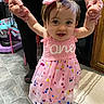 toddler, child, pink_dress, headband, bow, holding_hands, adult, kitchen, floor_tiles, rug, walking, happy, indoor, person, footwear, confetti_pattern, smiling, baby, celebration, assistance