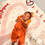 Reign joined the competition — help win amazing prizes! baby, infant, orange_outfit, headband, blanket, numbers, heart, cute, lying_down, indoors, soft_texture, curious_expression, barefoot, child, portrait, skin_tone, buttoned_clothing, playful, young_child, smiling