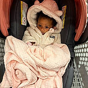 Reign is registered to the contest to win money with this photo: baby, child, car_seat, blanket, hoodie, pink, cream, face, eyes, infant, warm_clothing, cute, indoor, soft_texture, seat, baby_carrier, headwear, curious, wrapped, cozy