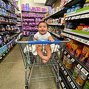 David is registered to the contest to win money with this photo: baby, shopping_cart, store, aisle, shelves, products, smiling, child, infant, happy, retail, shopping, floor, clothing, person, grocery, retail_shelf, container, package, indoor