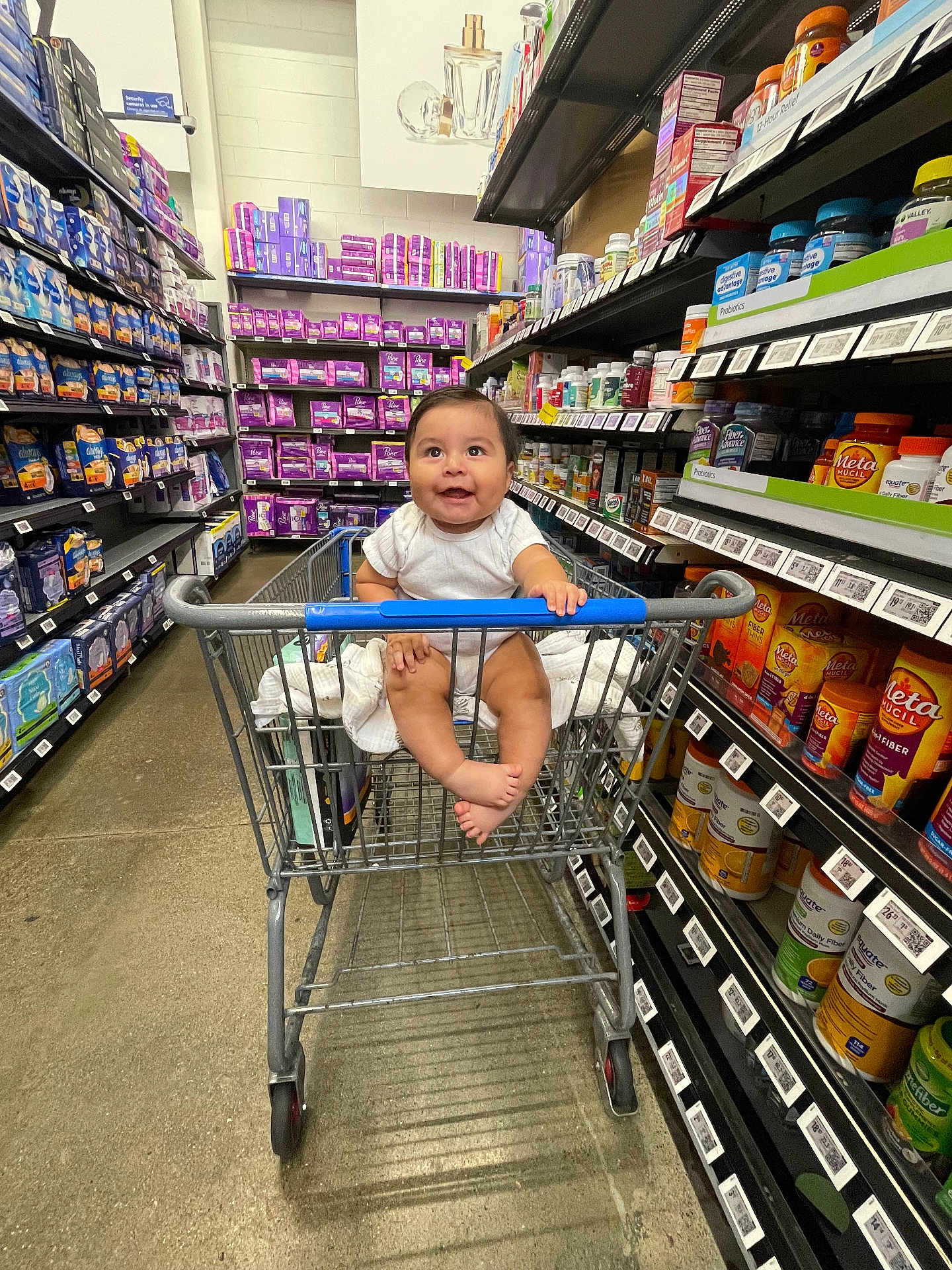 David is registered to the contest to win money with this photo: baby, shopping_cart, store, aisle, shelves, products, smiling, child, infant, happy, retail, shopping, floor, clothing, person, grocery, retail_shelf, container, package, indoor