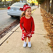 Ainara is registered to the contest to win money with this photo: child, sidewalk, red_clothing, hat, smile, curly_hair, white_shoes, car, street, sunset, trees, urban, outdoor, fence, concrete, happy, person, winter_wear, walking, daylight