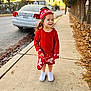 child, sidewalk, red_clothing, hat, smile, curly_hair, white_shoes, car, street, sunset, trees, urban, outdoor, fence, concrete, happy, person, winter_wear, walking, daylight