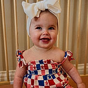 Hallie is registered to the contest to win money with this photo: baby, child, smiling, headband, bow, dress, red, white, blue, star_pattern, sitting, indoors, face, happy, cute, person, infant, wooden_baby_gate, skin, portrait