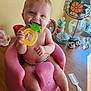 baby, infant, teether, pink_seat, wooden_table, lamp, stained_glass, decorative_items, glass_containers, photo_frame, cow_figurines, comb, smiling, chewing, cute, indoors, child, toy, happy, person