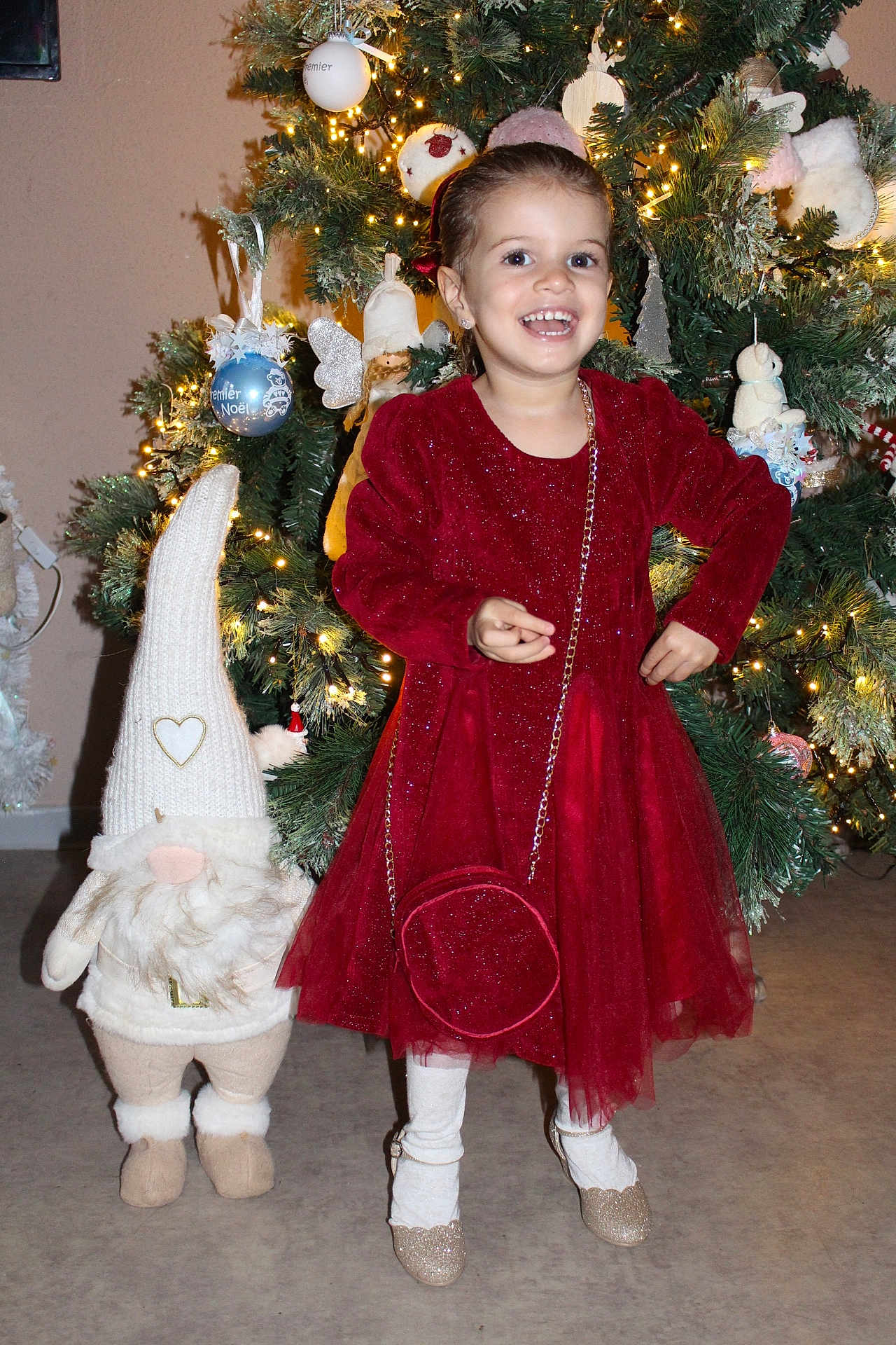 Tiana participe au concours pour gagner de l'argent avec cette photo : child, girl, red_dress, christmas_tree, holiday, festive, ornaments, lights, smile, happy, indoor, decorations, plush_toy, gnome, white_tights, shoes, pose, celebration, seasonal, joy