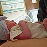 arm, baby, bed, blanket, care, child, clothing, hat, healthcare, holding, hospital, indoor, infant, medical_equipment, newborn, person, poster, resting, room, sleeping
