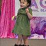 toddler, child, dress, green_dress, hat, curly_hair, boots, sparkly_boots, stage, pink_curtain, background_banner, posing, smiling, indoor, fashion, person, footwear, cute, happy, event